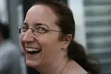 Photograph of Jeni Tennison. She is looking back over her shoulder toward the camera laughing. She is a white female with long dark hair, tied back, and is wearing glasses.