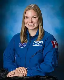 Jenni Sidey-Gibbons, wearing her blue Canadian Space Agency uniform bearing the flag of Canada, smiles at the camera, her hands folded.