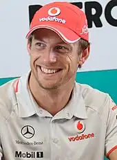 Jenson Button wearing a red baseball cap and silver T-short and smiling to the right of the camera