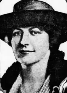 A white woman wearing a flat-brimmed hat, a fur coat or stole, and pearls