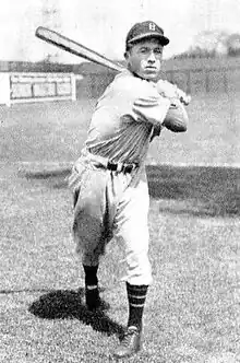 A man in a light a baseball uniform and dark cap posed as if having just swung his baseball bat