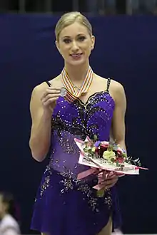 A woman in a blue dress smiles as she holds a medal and bouquet of flowers.