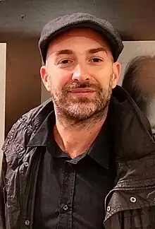 Color photograph of author Joel Willans standing next to a portrait of himself at a book fair booth