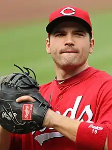 A man wearing a red baseball cap with a white "C" in the centre, a red baseball uniform with the words "REDS" written across but obscured by his left arm and a black baseball glove on his left hand with the words "RAWLINGS" sewn on it prepares to throw a baseball.