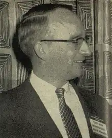 A man in a suit and tie. He is wearing a name badge on which can be discerned the words "Elroy McCaw, Washington State Assn. (Association) of Broadcasters".