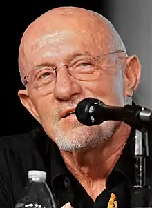 A bald middle-aged man wearing a black shirt, facing the camera, speaks into a microphone.