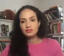 Duffy Rice is looking into the camera, and is seated in front of a bookcase. She has long curly hair, tan skin, and is wearing a pink crewneck sweater the same color as her lipstick.