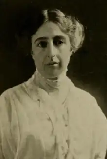 A middle-aged white woman, hair center-parted and dressed back from face and shoulders, wearing a high-collared white blouse or dress with ruffles