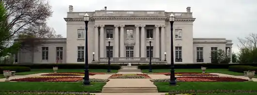 A two-story stone mansion with a sprawling flower garden in front