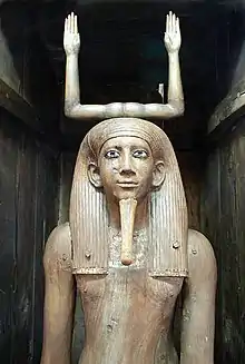 A picture of the top part of a wooden figure of a man with long hair. Two raised arms extend out of his head.