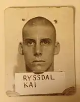 Sepia portrait of a man with a buzzcut holding up a card that says "RYSSDAL, KAI" on it