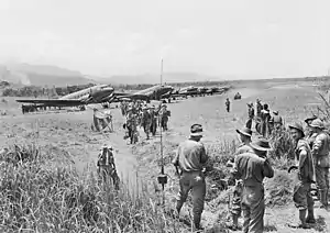 A line of Dakotas on a grass airstrip. Men wearing slouch hats file off a plane. A jeep drives along the strip. Other men in uniform and civilians look on.