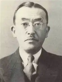 Close up of the head and shoulders of a solemn middle-aged Japanese man with a small mustache. He is wearing a suit, tie, and glasses.