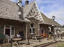 At bottom right is visible a rusted railway track and weeds growing between some of its ties. In the upper right is a cloudy sky, and the remainder of the photo is dominated by the pitched roof train station building, shrinking by perspective from left to right. There are two white-framed tripartite windows, each with a red door at its centre, flaking a central arch window underneath a protruding gable. In white lettering following the curve of the arch window is the text "Kensington". The wall is composed of rough stones of varying sizes. Between the rail and building is a boardwalk patio edged by a chain metal rail and dotted with numerous objects, including a wooden barrel, a picnic table, several smaller tables, stacked and unstacked chairs, and a lamppost from which hangs a flower basket.