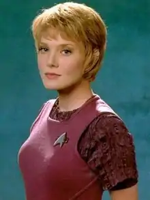 A photograph of a young woman with short blonde hair wearing a maroon outfit, with a Star Trek pin attached.