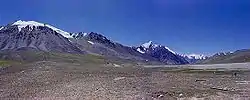 Some tall, bluegrey mountains rise out of brown soil below a deep blue sky