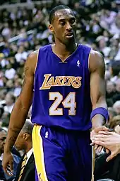 A man, wearing a purple jersey with a word "LAKERS" and the number "24" written in the front, is standing in front of the crowd.