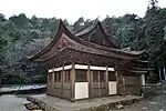 Small wooden building with white walls and hip-and-gable roof connected to another wooden structure in the back.