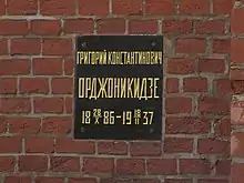 A black plaque with gold letters on a brick wall with a Russian inscription that translates as Grigory Konstantinovich Ordzhonikidze, 1886-10-28–1937-02-18