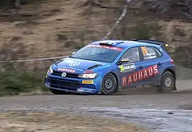  Five-door two-box car in a racing livery fitted with a rear aerofoil and a roof-mounted air scoop for ventilation travelling at speed on a gravel course