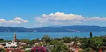 Buildings in a Spanish architectural style amid trees, with a blue body of water behind them and mountains and blue sky in the background