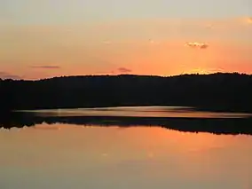 A red-orange sunset and the black silhouette of a line of trees are reflected in a smooth lake