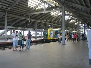 A relatively empty boarding platform with only a handful of people identified by a sign as that at Vito Cruz station. Pebbles line the tracks and sunlight comes in from spaces open to the outside and large open flaps in the dark warehouse-like roof.