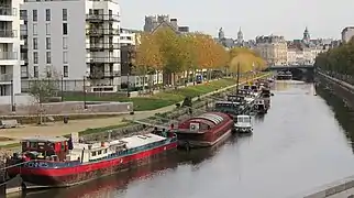 an urban river flowing past modern apartment buildings in the foreground past trees whose leaves are changing with autumn under a bridge and into the city center of Rennes. There are boats docked along the side of the river.