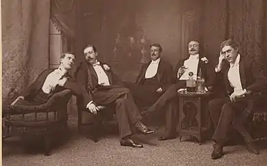 indoor stage scene showing five men relaxing in Victorian evening clothes