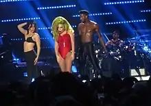 In the center of the photograph is a woman with blonde hair in red clothing; to her sides are two scantily-clad people, and in the background is someone sitting at a drum set.