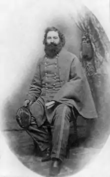 Black and white photo shows a seated man with a bushy beard. He wears a gray military uniform with two rows of buttons and a double pinstripe on the trousers.