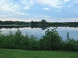 A placid lake lies under a pink/blue afternoon sky. In the foreground is a well groomed lawn and a border of plants, including a sunflower, that encircles that lake.