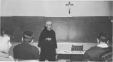 Three young men listen as an elderly monk gives a lecture in front of a chalkboard, a desk with a chair, and a crucifix.