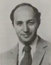 A black and white portrait of a man with thinning hair, wearing a suit and tie, facing the camera while grinning.