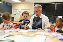 Three children and one adult are sitting and painting at a classroom table.