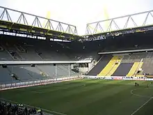 Two-tiered stands with grey seats, below the stands is a pitch with white markings