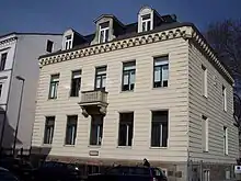  Light-coloured two-storey stone building with an ornamental ledge below the roof. There are rows of five windows at ground level and the floor above, with a balcony at the middle upper window.