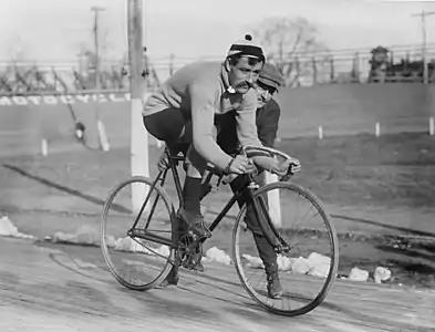 A black-and-white picture of a man on an old bicycle. Another man is holding or pushing the bicycle.