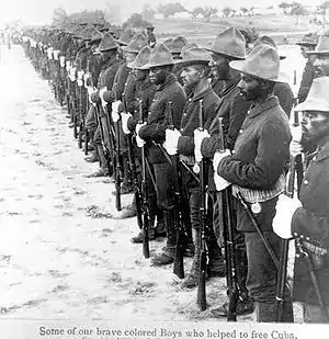 A black and white photo of the liberators of Cuba. The picture shows a line of African-American Buffalo Soldiers in a line with rifles and equipment with the right foreground the closest part of the line. The line extends into the left background. The soldiers are standing tall looking to the left of the picture.