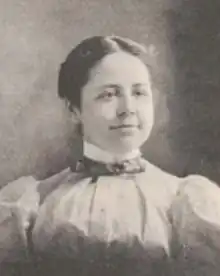 A young white woman with dark hair pared center and dressed back to the nape, wearing a high-collared blouse with puffy sleeves and a bow-tie at the throat