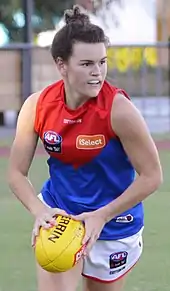 A female athlete in a red and blue sleeveless guernsey running with a football