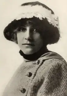 A white woman wearing a hat with a brim and feather embellishment; she is also wearing a coat with an off-center buttoned closure