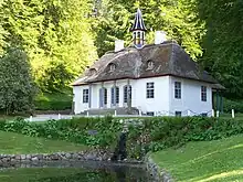 A thatched and plaster country house sits on a rise with trees in the background. The roof has three gables and a columned cupola with a pointed top. Steps go up to the three arched paned glass entrances on the columned porch.