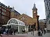 The end of a brick building with arched windows and sloping roofs lies between two towers with steeples. In front of this is a white metal and glass structure. People are standing and walking in the street in front.