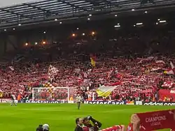 A single-tiered stand that contains thousands of people. Several flags are being waved. In front of the stand is a grass pitch with a goal.