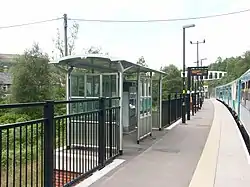 Covered waiting area of metal and glass, on mortared brick support extended from platform. Woods behind railings to left of platform edge. Four carriage train to the other.