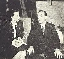 A dark-haired woman siting with a notebook in her lap smiles at a man in a suit and tie as he speaks.