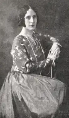 A white woman with dark marcelled hair, seated, wearing a dress with a print bodice; her hands and clasped together in front of her chest