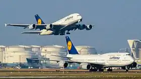 A Lufthansa Airbus A380 in the air about to land. In the foreground, a Lufthansa Boeing 747-8, is on the ground taxiing on a taxiway.