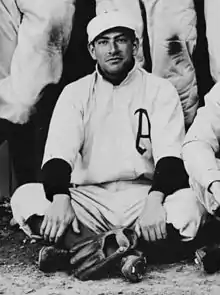 A man sitting on the ground wearing a white baseball uniform with an "A" on the chest and a baseball glove placed at his feet